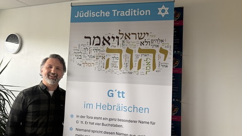 Mustafa Cimşit steht vor einer Plakatwand für eine Wanderausstellung von Maimonides für Schulen im Kampf gegen den Antisemitismus. 