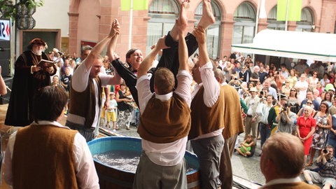 Eine junge Frau wird beim Johannisfest gegautscht. Sie wird mehrmals in einen Bottich mit Wasser getaucht. 