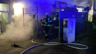 Feuerwehrleute kontrollieren bei Nacht gelöschte, noch rauchende Mülltonnen neben einem Einkaufsmarkt.