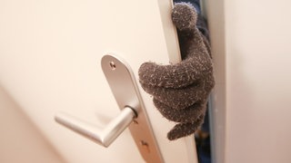 Eine Person, die Stoffhandschuhe trägt, betritt eine fremde Wohnung. Die Mainer Polizei warnt vor einer Betrugsmasche. 