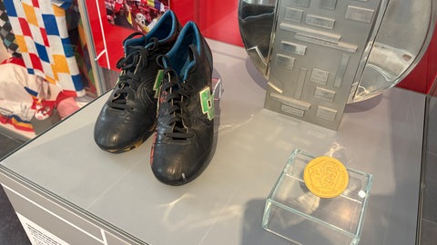 Fußballschuhe stehen neben einer Medaille in einer Vitrine.