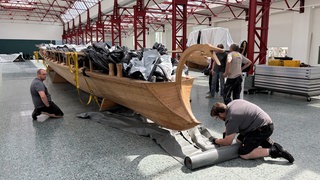 Männer einer Spezialfirma legen eine Plastik-Plane unter ein Schiff, das verschoben wird. 
