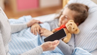 Eine Mutter sitzt mit Handy in der Hand am Bett ihres kranken Kindes.