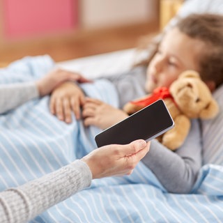 Eine Mutter sitzt mit Handy in der Hand am Bett ihres kranken Kindes.