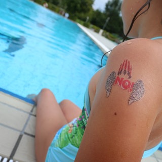 Ein Mädchen mit einem "NO!" Klebetattoo am Arm sitzt im Freibad am Beckenrand. 