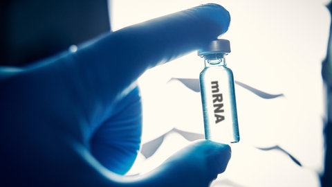 Eine Hand mit blauem Gummihandschuh hält ein Glasfläschen mit der Aufschrift mRNA.
