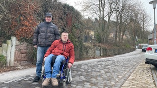 Rollstuhlfahrer Patrick Loppnow und Michael Scheiwen stehen vor der steilen Straße die zur Egbert-Grundschule in Trier-Kürenz führt. Den Weg zum Wahllokal finden sie nicht barrierefrei.