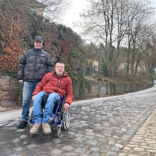 Rollstuhlfahrer Patrick Loppnow und Michael Scheiwen stehen vor der steilen Straße die zur Egbert-Grundschule in Trier-Kürenz führt. Den Weg zum Wahllokal finden sie nicht barrierefrei.