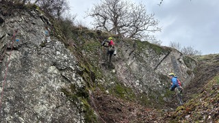 Im Moment klettern noch Arbeiter an Arbeitsseilen in der Felswand. Bald soll hier jeder auf dem neuen Klettersteig in Neuerburg klettern können, der darauf Lust hat, eine spezielle Ausrüstung nutzt, und eingewiesen wurde.