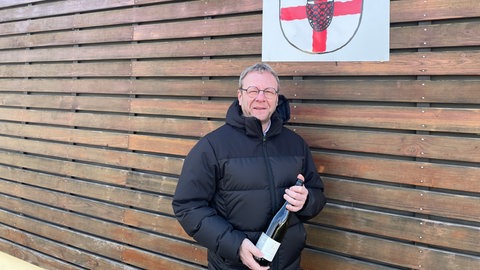 Der Piesporter Ortsbürgermeister steht mit einer Weinflasche in der Hand vor der Tourist-Information.