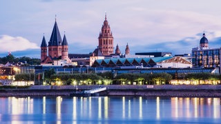 Stadtansicht von Mainz im blauen Abendlicht, im Vordergrund der Rhein, im Hintergrund der Dom, die Rheingoldhalle und die Kirche St. Stephan.