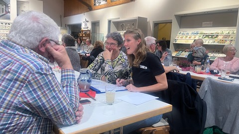 Drei Menschen sitzen an einem Tisch und unterhalten sich, die beiden Frauen sind der Kamera zugewandt, der Mann sitzt mit dem Rücken zur Kamera.