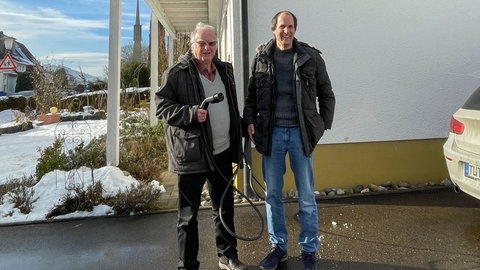 Gustav Schlecht mit Sohn Harald Schlecht vor ihrem Haus, neben einem Auto. Gustav Schlecht hat ein Ladekabel für ein E-Auto in der Hand.