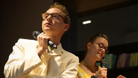 Schauspieler Carl Grübel bei Liederabend, im Hintergrund Schauspielerin mit gelbem Kleid