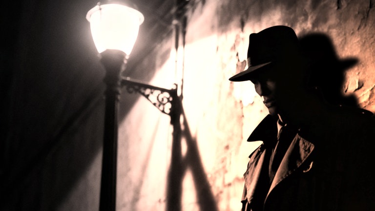Leo Malet: Das Leben ist zum Kotzen - Ein Mann mittleren Alters steht an eine Mauer gelehnt im Schein einer alten Straßenlaterne. Er trägt einen Hut und einen Trenchcoat, sein Gesicht bleibt im Schatten verborgen. 