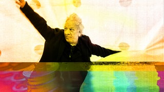 Der Autor, Komponist und Musiker FM Einheit breitet die Arme aus vor einem geschwungenen gelben Hintergrund, unter seinen Füßen sieht man links Maschinenteile, lila eingefärbt, und rechts bunte Farbverläufe.