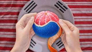 Zwei Hände schälen eine Mandarine, deren Fruchtfleisch und Schale in den Farben Rot, Weiß und Blau gestaltet sind. Ein Stück der orangefarbenen Schale hängt nach unten. Im Hintergrund ist eine graue Fläche zu sehen, überlagert von Elementen der südkoreanischen Flagge mit schwarzen Trigrammen und roten Bereichen.