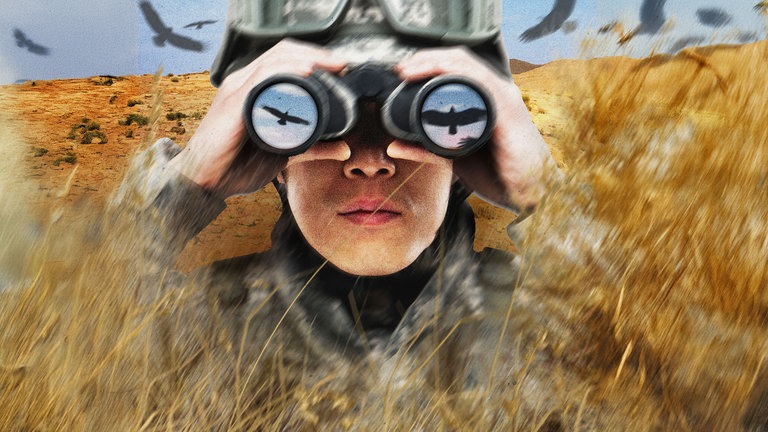 Ein US-amerikanischer Soldat beobachtet durch sein Fernglas Falken inmitten einer Wüstenlandschaft.