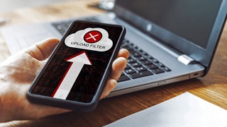 Ein Smartphone wird von einer Hand vor einem Laptop gehalten. Auf dem Bildschirm des Smartphones ist ein Cloud-Symbol mit einem roten X angezeigt, sowie ein Pfeil, der in die Cloud weist. Außerdem steht unter der Cloud „Upload-Filter“.