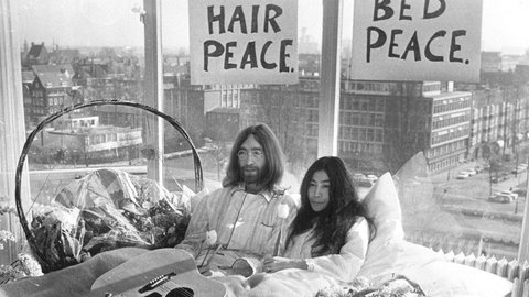 John Lennon (links) und Yoko Ono sitzen in einem Bett, im Hintergrund ist Amsterdam zu sehen. 
