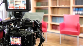 Zwei leere Stühle in einem Fernsehstudio mit einem Bücherregal im Hintergrund und einer Kamera im Vordergrund