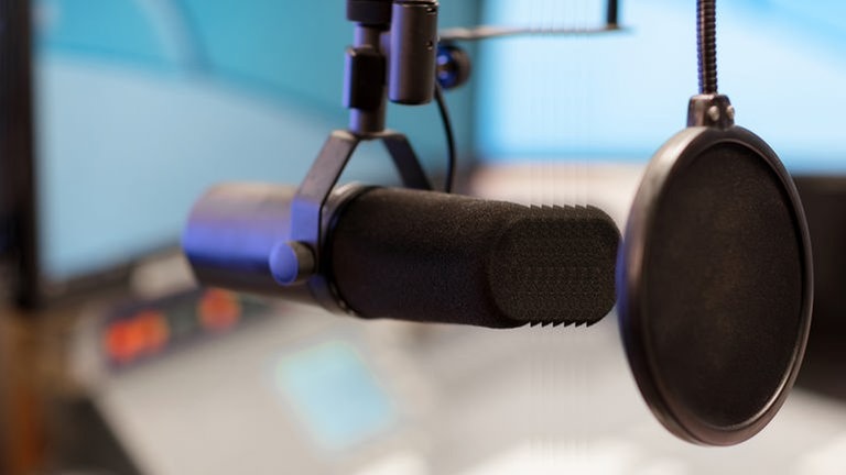 Ein Mikrofon mit Ploppschutz in einem Studio, im Hintergrund ein Mischpult mit Monitor