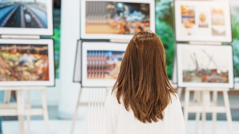 Eine Frau mit braunen langen Haaren steht vor mehreren Staffeleien mit Bilder