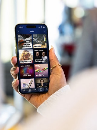 Eine Person mit Kopfhörern schaut auf ein Smartphone, auf dem das Angebot von SWR Kultur in ARD Sounds zu sehen ist