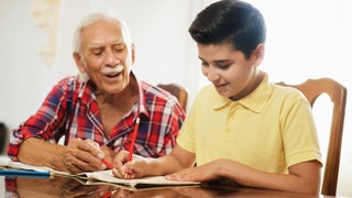 Ein älterer Mann hilft einem Jungen bei den Hausaufgaben: Persönliche Mentorinnen oder Mentoren sollen Kindern aus sogenannten bildungsfernen Schichten helfen, ihre Ziele zu erreichen. Die Angebote reichen von Hausaufgabenhilfe bis zu Ausflügen in den Zoo oder ins Museum.