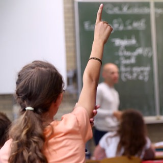 Ein Mädchen streckt im Unterricht den Finger, ein Lehrer erklärt an der Tafel: Demokratiebildung ist Aufgabe von Schule. Der sogenannte "Beutelsbacher Konsens" aus dem Jahr 1976 legt die Prinzipien dafür fest: Keine Indoktrination im Klassenzimmer, politische Kontroversen sind auch kontrovers darzustellen, zudem sollen Schüler*innen befähigt werden, sich eine eigene Meinung zu bilden. 