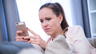 Junge Frau schaut mit verzogener Miene auf ihr Smartphone: Häufig sind es Frauen, die Opfer von Cyberstalking werden. Sie werden beleidigt und in Kommentaren und Videos auf sozialen Medien bedroht. Vielen setzt das psychisch stark zu. 