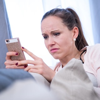 Junge Frau schaut mit verzogener Miene auf ihr Smartphone: Häufig sind es Frauen, die Opfer von Cyberstalking werden. Sie werden beleidigt und in Kommentaren und Videos auf sozialen Medien bedroht. Vielen setzt das psychisch stark zu. 