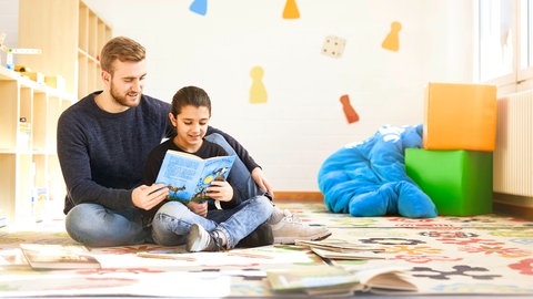 Ein Junge und ein Student sitzen im Schneidersitz auf dem Boden und üben lesen: Rund 1.200 Mädchen und Jungen wurden 2021 durch die „Kinderhelden“ gefördert – nicht nur beim Lesen, sondern auch in Mathematik oder den Naturwissenschaften.