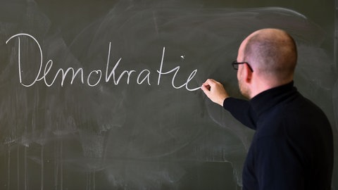 Ein Lehrer schreibt mit Kreide das Wort "Demokratie" an eine Schultafel: Kinder und Jugendliche sollten möglichst früh ihre Rechte, Pflichten und ihren Selbstwert in der Gesellschaft kennen: teilhaben, mitgestalten und Eigenverantwortung übernehmen von Anfang an