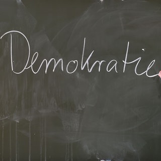 Ein Lehrer schreibt mit Kreide das Wort "Demokratie" an eine Schultafel: Kinder und Jugendliche sollten möglichst früh ihre Rechte, Pflichten und ihren Selbstwert in der Gesellschaft kennen: teilhaben, mitgestalten und Eigenverantwortung übernehmen von Anfang an