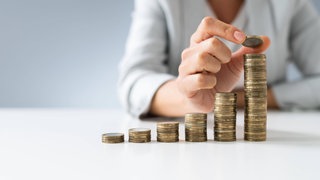 Eine Frau stapelt Geldmünzen vor sich: Frauen sparen weniger, kaufen seltener Aktien oder investieren in ETFs. Und sie sorgen nicht genug fürs Alter vor. Die Folge: Oft sind sie finanziell abhängig von (Ehe-)Männern und ihnen droht Altersarmut.