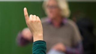 Schüler streckt Finger, im Hintergrund ein Lehrer: An vielen Schulen herrscht Lehrermangel. Fachkräfte, die kein Lehramt studiert haben, füllen die Lücken und unterrichten Mathe, Chemie oder Musik.