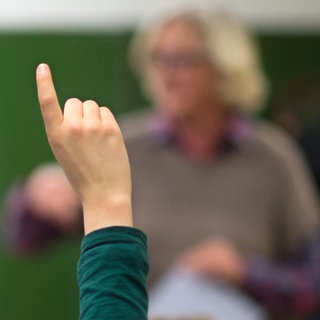 Schüler streckt Finger, im Hintergrund ein Lehrer: An vielen Schulen herrscht Lehrermangel. Fachkräfte, die kein Lehramt studiert haben, füllen die Lücken und unterrichten Mathe, Chemie oder Musik.
