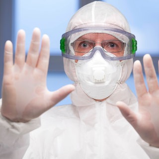 Eine Person im Schutzanzug hebt abwehrend die Hände: Früher oder später wird sich wieder eine neuartige Infektion global ausbreiten, darüber sind sich Fachleute aus Medizin und Gesundheitspolitik einig. Wie können sich Staaten vorbereiten und wie lassen sich Fehler verhindern?