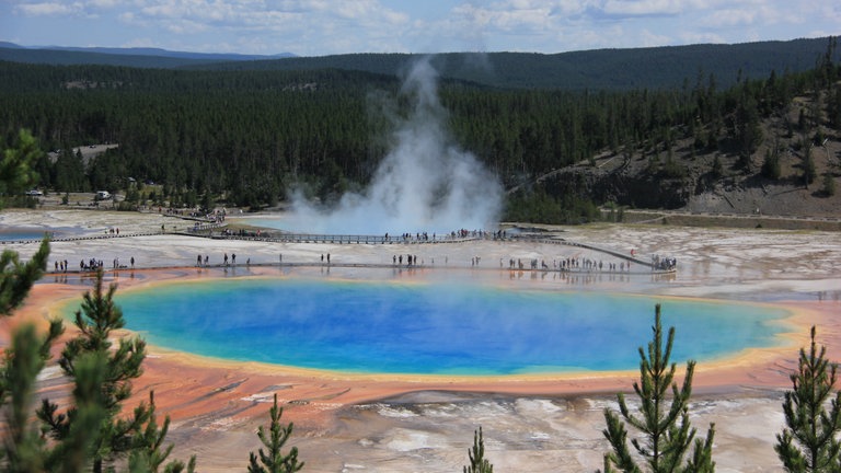 Ein blauer See inmitten von Berglandschaft: Für Schlagzeilen sorgen jeden Sommer Touristen, die sich Tieren zu sehr nähern oder in heiße Quellen springen, in denen Mikroorganismen abhängig von der Temperatur den Grund strahlend Türkis, Rot und Geld färben – hier der „Grand Prismatic Spring“