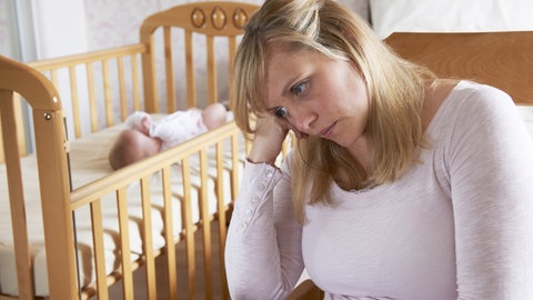 Mutter sitzt traurig vor einem Bettchen, in dem das Baby schläft: Nach der Geburt erleben viele Mütter einen Babyblues. Bleibt dieses Gefühl, haben sie eine Wochenbettdepression. Die Erkrankung kann jede Mutter treffen, bleibt aber oft unerkannt.