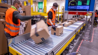 Zwei Männer legen Pakete auf ein Band im Verteilzentrum des Online-Händlers Amazon in Mecklenburg-Vorpommern: Der Anteil von Amazon am Online-Markt liegt in Deutschland bei 55 Prozent, in den USA bei über 70. Missbraucht der Konzern seine Marktmacht zum Nachteil des Wettbewerbs und diktiert die Preise?