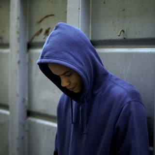 Junge mit blauem Kapuzenshirt lehnt an einem Gitter und schaut nach unten: Etwa 40.000 Minderjährige und junge Menschen in Deutschland haben kein festes Zuhause. Was treibt die Jungendlichen auf die Straße? Wann hilft ihnen der Staat, wo sind Lücken? Und was wünschen sich junge Obdachlose?