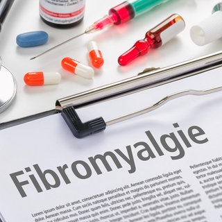 Klemmbrett mit einem Infoblatt zur Fibromyalgie, dazu Medikamente und ärztliche Instrumente:Wer an Fibromyalgie leidet, hat oft starke Schmerzen. Meist trifft es Frauen. Lange galt die Erkrankung als psychosomatisch. Dabei sind die Ursachen komplex – und noch unklar.