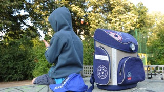 Ein Kind im Kapuzenpulli sitzt mit seinem Schulranzen im Park: Die Gründe für Schulverweigerung sind vielfältig: Häufig haben die Kinder und Jugendlichen Angst vor der Schule, weil sie Mobbing, Stress durch Leistungsdruck oder Probleme mit Lehrkräften erlebt haben. 