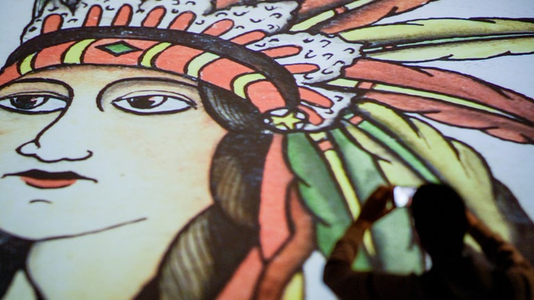 Kopf einer Indianerin mit Federschmuck: Handgemalten Vorlagezeichnungen mit ihrer Vielfalt an Motiven waren 2019 Teil der Ausstellung "Tattoo-Legenden. Christian Warlich auf St. Pauli" im Museum für Hamburgische Geschichte. Hier fotografiert ein Besucher das Exponat