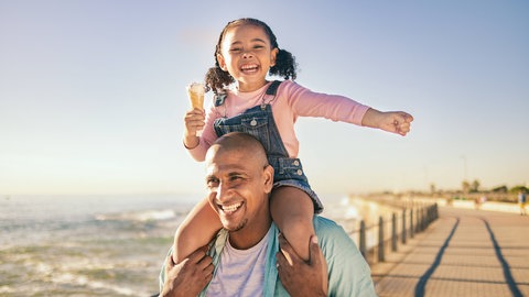 Kleines Mädchen mit einem Eis in der Hand sitzt glücklich lachend auf den Schultern des Vaters: In der Erziehung sind auch Ausnahmen von Regeln erlaubt: Ein Extra-Eis sorgt nicht dafür, dass das Kind den Eltern auf der Nase herumtanzt.