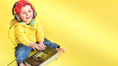 Kleinkind mit roter Mütze hört Radio über Kopfhörer: Noch ist das Radio der Zukunft Spekulation. Denkbar ist eine gemeinsame Plattform mit der Möglichkeit, unter verschiedenen Anbietern auszuwählen – das, was heute die Radiosender sind. Die Plattform kann über verschiedene Endgeräte empfangen werden: Smartphones, Computer, aber auch klassisch über das Auto- oder Küchenradio. 