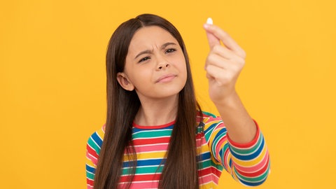 Mädchen hält skeptisch ein Dragee in die Höhe: Kinder brauchen für ihre gesunde Entwicklung Vitamine und Mineralstoffe. Aber was, wenn sie sie nicht über die Nahrung aufnehmen können oder wollen? Dann gibt es Nahrungsergänzungsmittel. Aber bei welchen Kindern ist die Nahrungsergänzung wirklich sinnvoll und welche Eltern sollten lieber die Finger davon lassen?