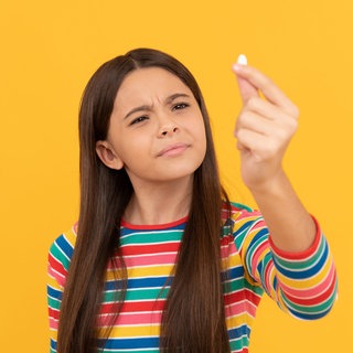 Mädchen hält skeptisch ein Dragee in die Höhe: Kinder brauchen für ihre gesunde Entwicklung Vitamine und Mineralstoffe. Aber was, wenn sie sie nicht über die Nahrung aufnehmen können oder wollen? Dann gibt es Nahrungsergänzungsmittel. Aber bei welchen Kindern ist die Nahrungsergänzung wirklich sinnvoll und welche Eltern sollten lieber die Finger davon lassen?